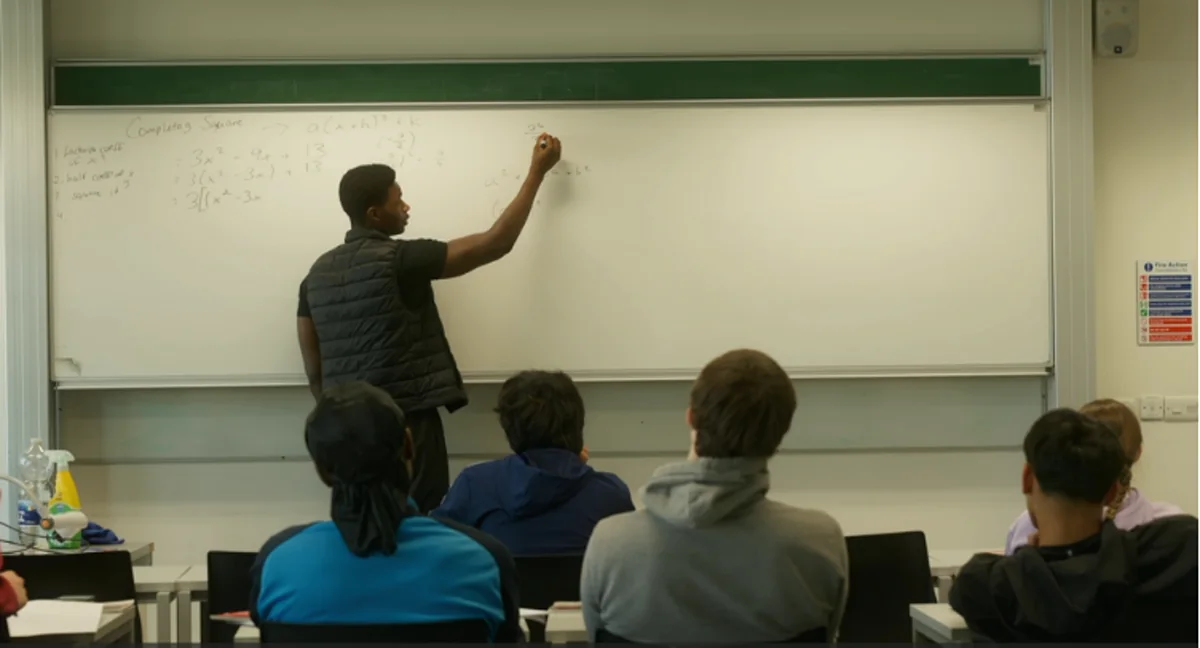 A tutor working through completing the square on a whiteboard in front of a class of students in a lecture room
